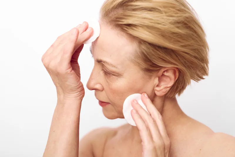 Mature woman with short hair gently cleanses face with cotton pads on white background. Concept of skincare, aging skin care, and daily beauty routine for mature skin.