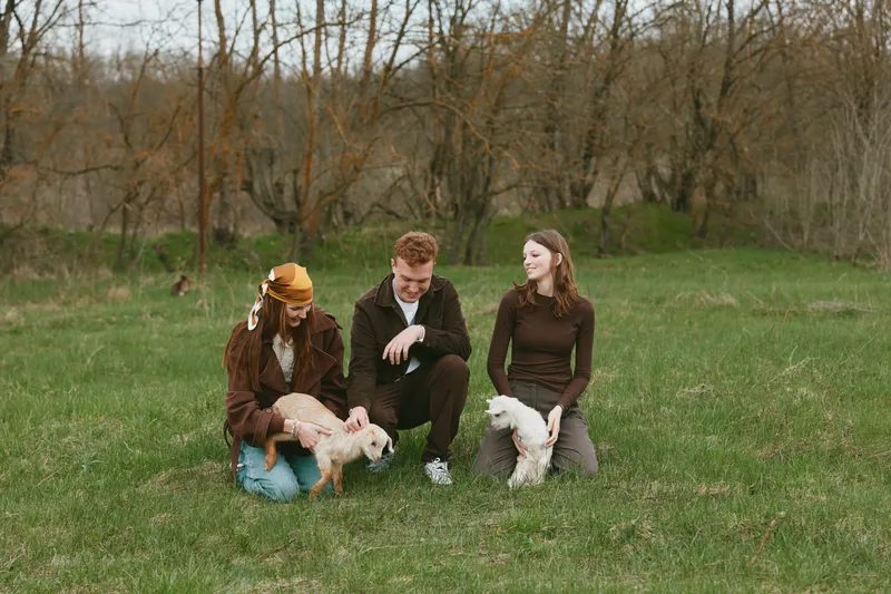 Three people in a grassy park with two small dogs, casual autumn outfits, candid lifestyle moment, friendship and companionship, relaxed conversation, natural outdoor lighting, wholesome mood.