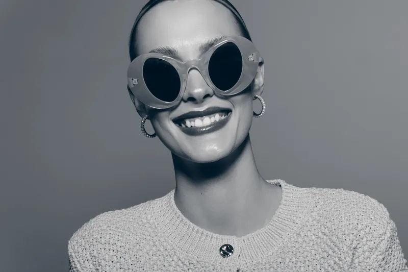 Smiling Caucasian woman with pale skin wearing round sunglasses and hoop earrings in knitted top. Bright joyful expression on grey background for fashion and lifestyle concept.