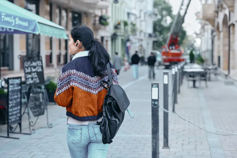 Woman with backpack walks along a city street wearing an orange patterned jacket and jeans, urban fashion in motion among cafe signs and pedestrian traffic