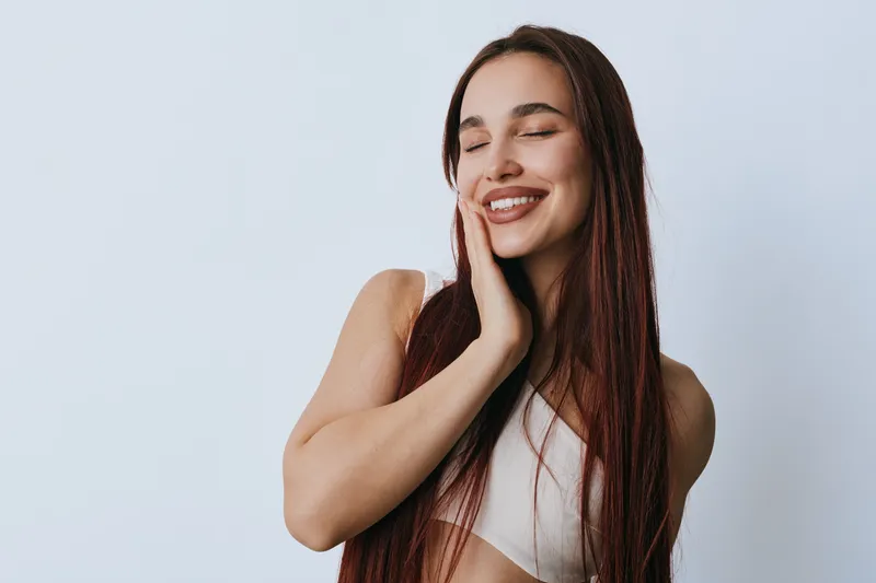Young woman with long hair smiling and touching face in white top on light background. Happy female with closed eyes and natural beauty in casual pose with relaxed expression.