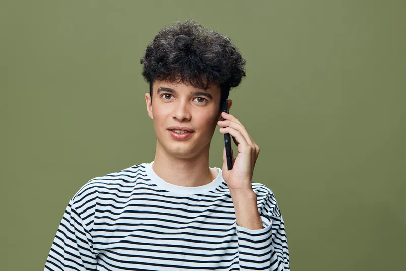 young man talking on smartphone in casual striped shirt, curly hair, olive green background, engaging expression, modern lifestyle concept