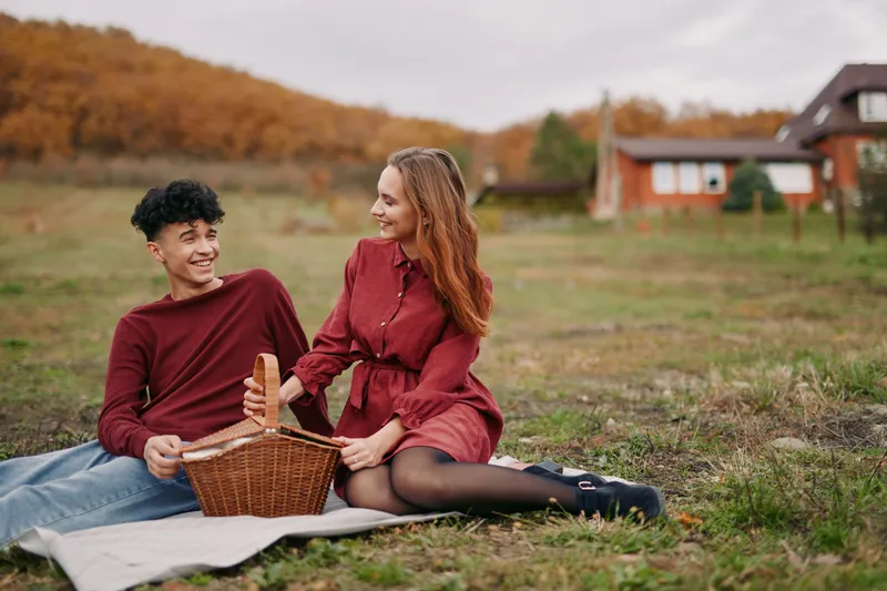 A genuine couple sharing a relaxed outdoor picnic on a grassy field, capturing authentic interaction and natural smiles as they enjoy a warm autumn afternoon together with a wicker basket,