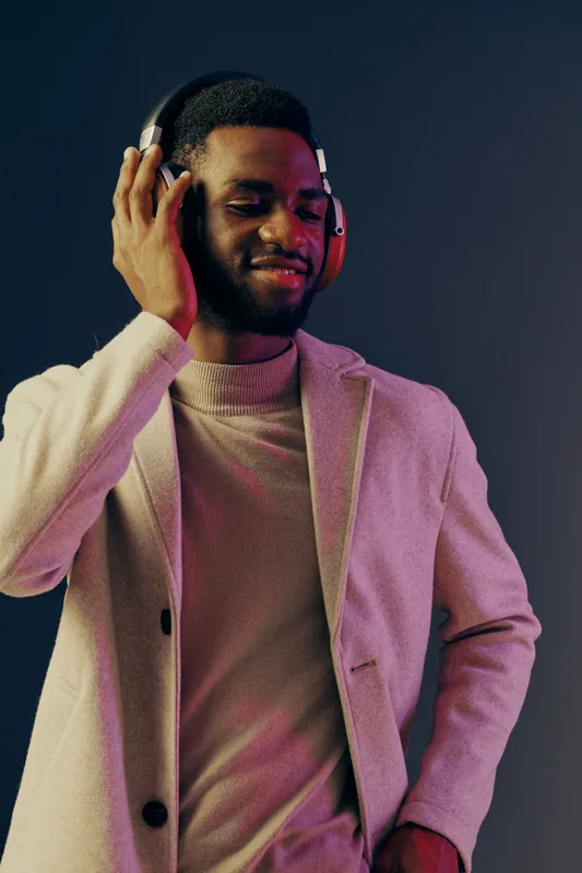A young African man enjoying music with headphones, dressed in stylish casual attire, set against a gradient background in soft pastel colors, portraying happiness