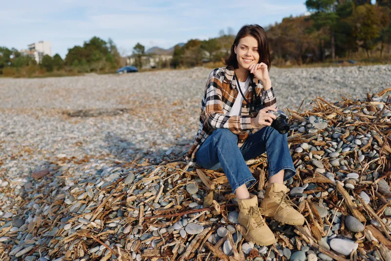 Woman, beach, shells, casual, outdoor, smile, sitting, coastal, portrait, lifestyle. A relaxed moment on a shell beach with jeans and plaid shirt under natural light, capturing a cheerful vibe and