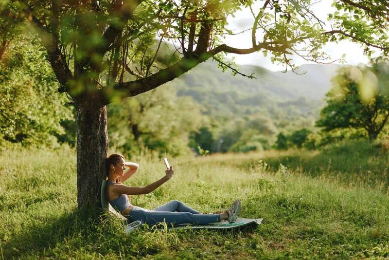 Young woman enjoying a sunny day in the park, taking a selfie under a tree, surrounded by vibrant green foliage, wearing casual denim outfit