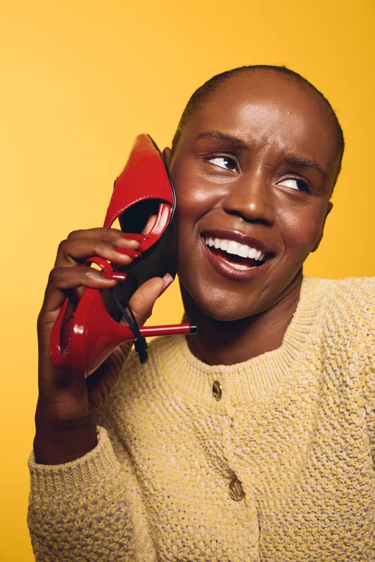 Happy African woman with dark skin smiles while holding a red high heel shoe to her ear on yellow background. Concept of fashion, joy, and playful lifestyle with bright colors.