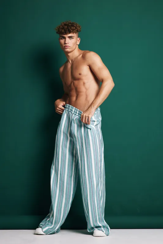 Muscular young man posing shirtless wearing loose striped pants standing against green wall studio background with serious expression. Model showcasing athletic body and curly hair fashion casual