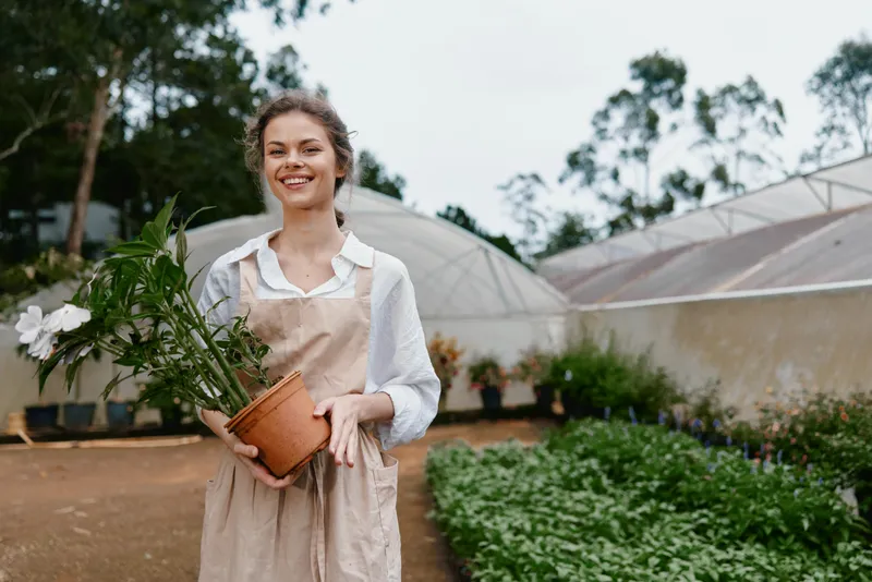 Smiling woman in an apron holding a potted plant in a greenhouse, embodying joy and connection to nature through gardening