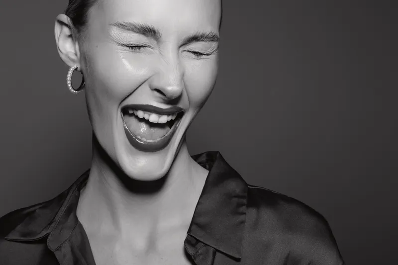 Young white woman with fair skin and short hair laughing with eyes closed, wearing hoop earrings and a satin blouse. Studio portrait in black and white. Concept of joy and happiness.