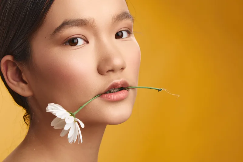 asian woman gently holds a delicate flower between her lips, looking softly at the camera in a bright studio, conveying calm elegance, natural beauty, and intimate minimalism.