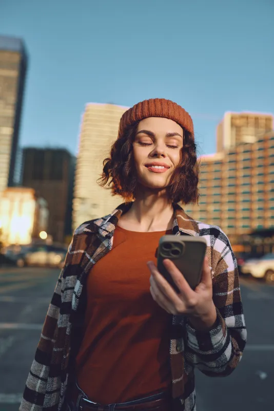 woman smartphone smile urban beanie plaid young portrait in golden hour glow, candid lifestyle moment with authenticity and emotional storytelling, relaxed city walker checking mobile.