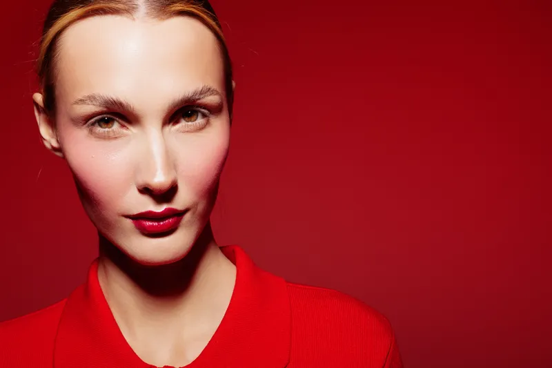 Close-up portrait of confident woman with red lips and red shirt on red background. Studio lighting highlights facial features and creates bold, striking mood for fashion concept.
