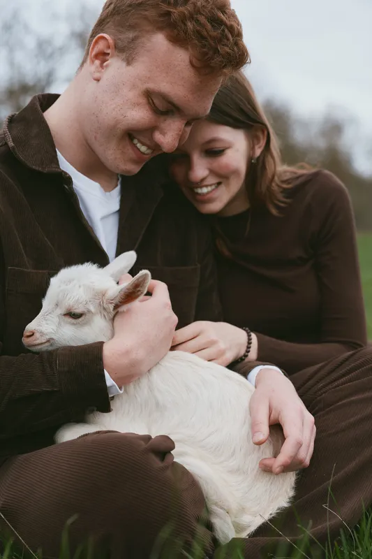 Happy couple outdoors cuddling a young goat kid in a grassy field, a cozy, affectionate moment highlighting farm life, connection, and gentle animal care in natural light.