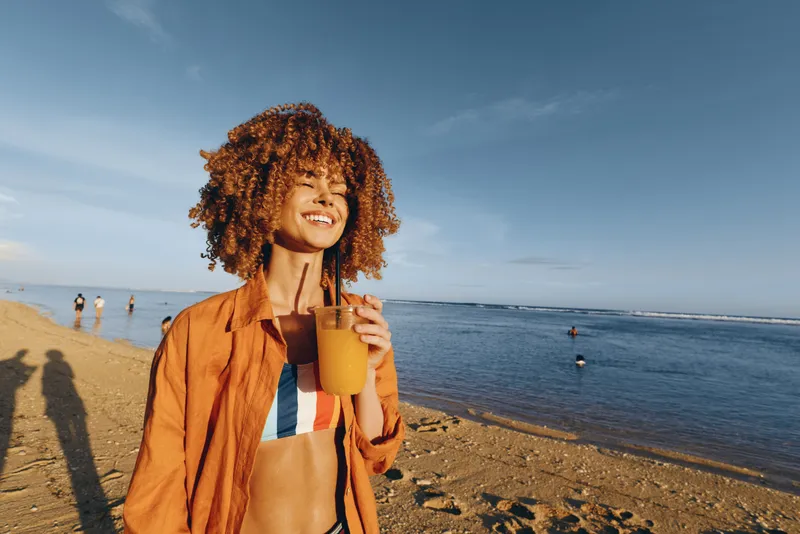 Smiling young woman with Afro curly hair on a sunny beach near the ocean, holding a coffee cup, wearing an orange jacket, relaxed vacation mood, clear blue sky horizon