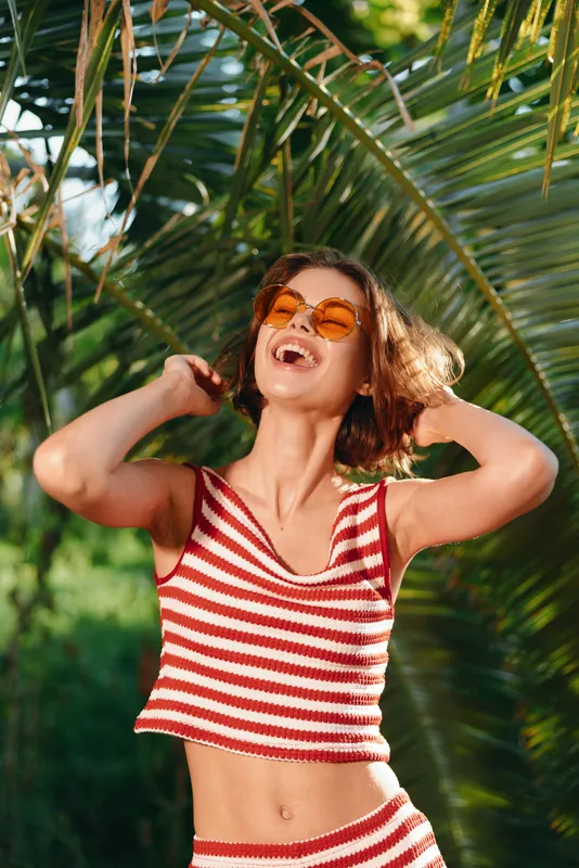 A cheerful woman wearing a red and white striped crop top and sunglasses basks in sunlight, smiling with arms raised, enjoying a carefree moment outdoors.