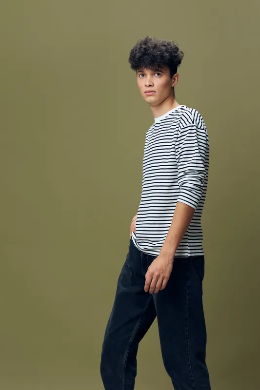 young man with curly hair wearing black and white striped long sleeve shirt and dark jeans stands against olive background, casual style and urban fashion concept.