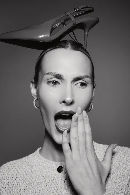 Young white woman with pale skin and slicked hair yawns covering mouth. High heel shoe balanced on head. Studio portrait in monochrome. Concept for fashion, fatigue, and style.