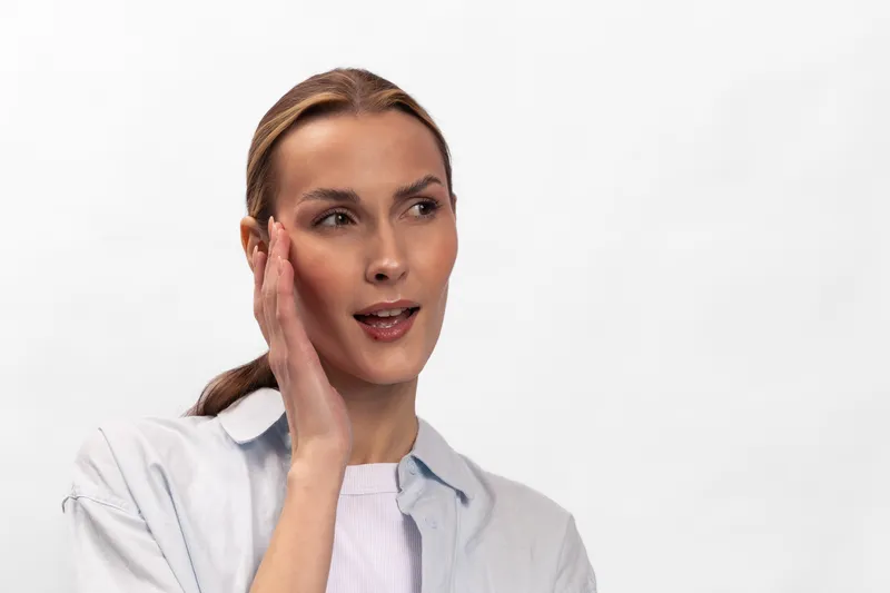 Young woman with light skin touching her face, looking thoughtful on white background. Concept of skincare, beauty routine, self-care, and natural facial expression in studio lighting.