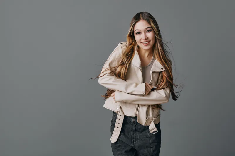 Young woman with long hair wearing beige jacket and black jeans smiling and posing with crossed arms in studio with gray background and casual style.