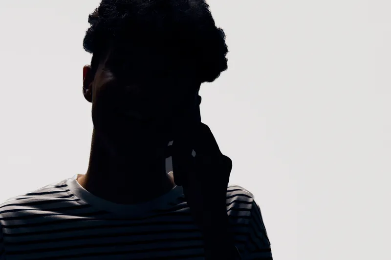 Silhouetted man wearing striped shirt holding phone, dark shadowed figure with indistinct facial features on light background, minimalistic monochrome contrast.