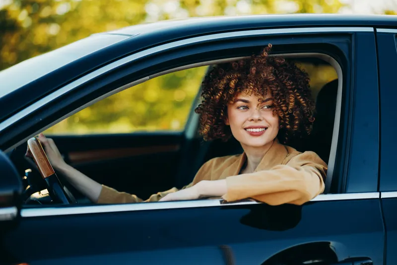 Young woman with curly hair sits in a blue car and smiles as she drives along a tree lined road with a retro film color mood, suggesting carefree daily life