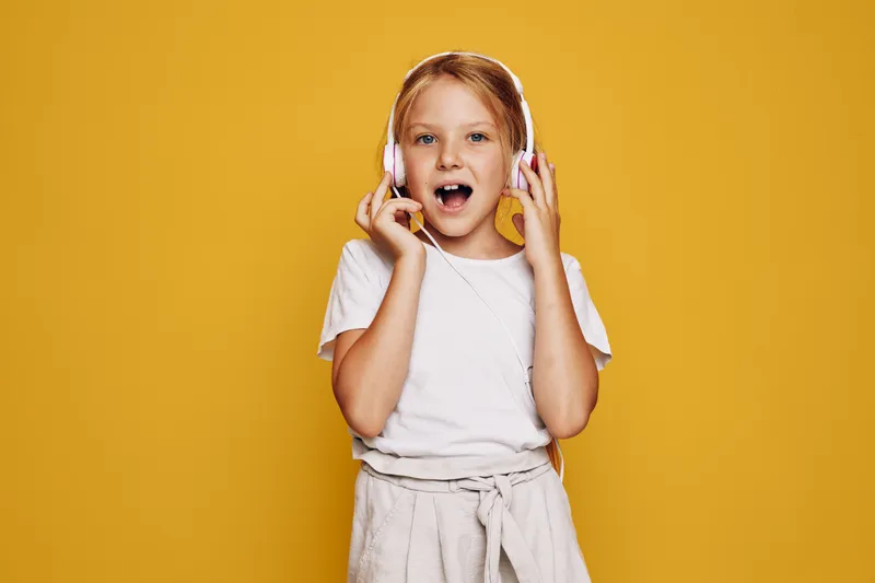 Young girl with blonde hair wearing headphones listens to music and smiles on a yellow background. Casual outfit with white shirt and beige pants.