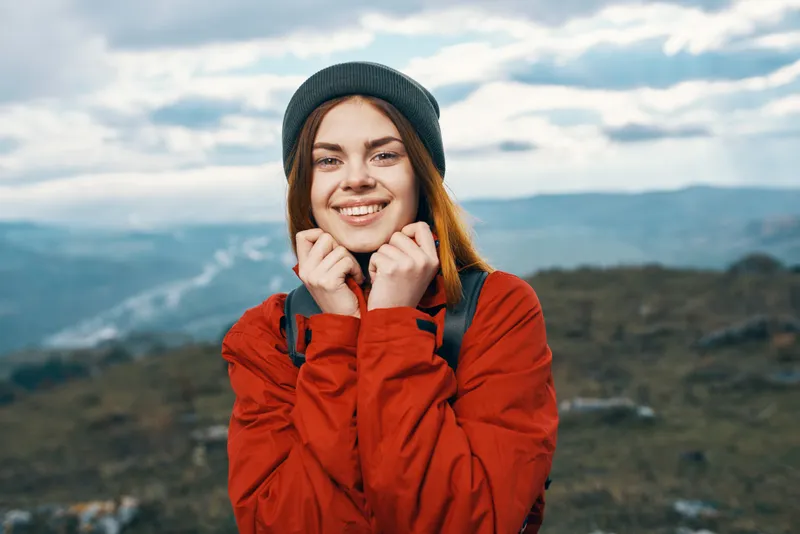 Счастливая женщина в красной куртке с рюкзаком и в шапке, на заднем плане горы, свежий воздух