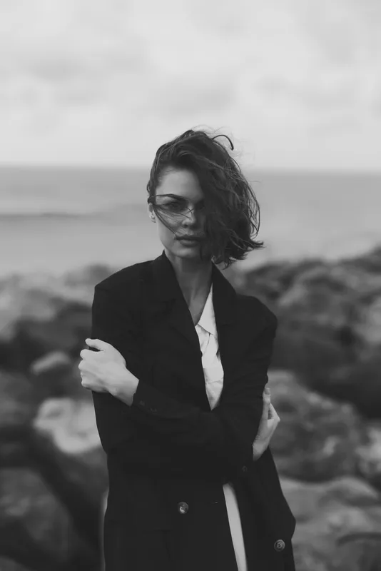 Woman in a dark coat stands by a rocky shore, wind sweeping hair, monochrome mood, fashion photography by the sea with an outdoor dramatic ocean backdrop