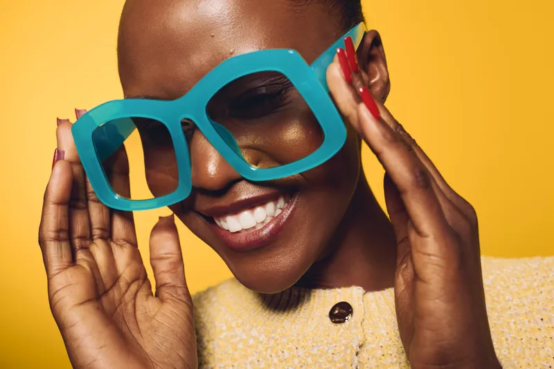 Smiling dark skinned African woman wearing oversized blue glasses and yellow top poses against yellow background. Concept of fashion, beauty, joy, and modern style for women.