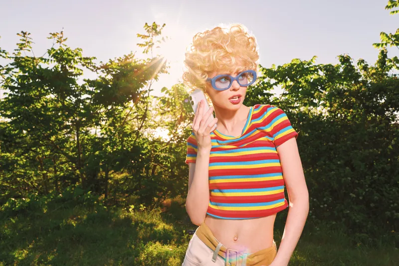 Vibrant retro woman outdoors wearing a striped crop top and sunglasses, curly blond wig, talking on a phone. Bright sun, green surroundings, playful lifestyle, warm nostalgic mood.
