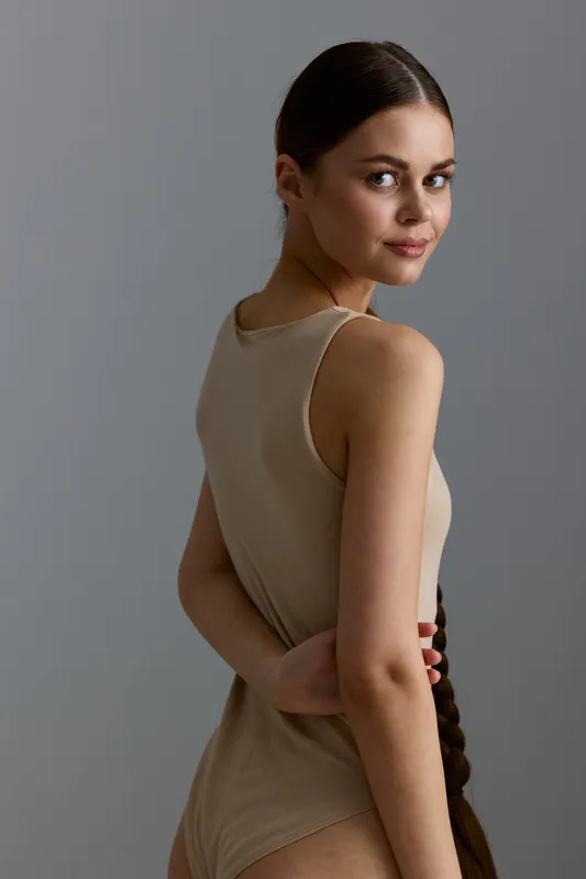 Young woman with long braided hair wearing a beige sleeveless bodysuit, posing against a plain grey background, showing a casual and natural look.