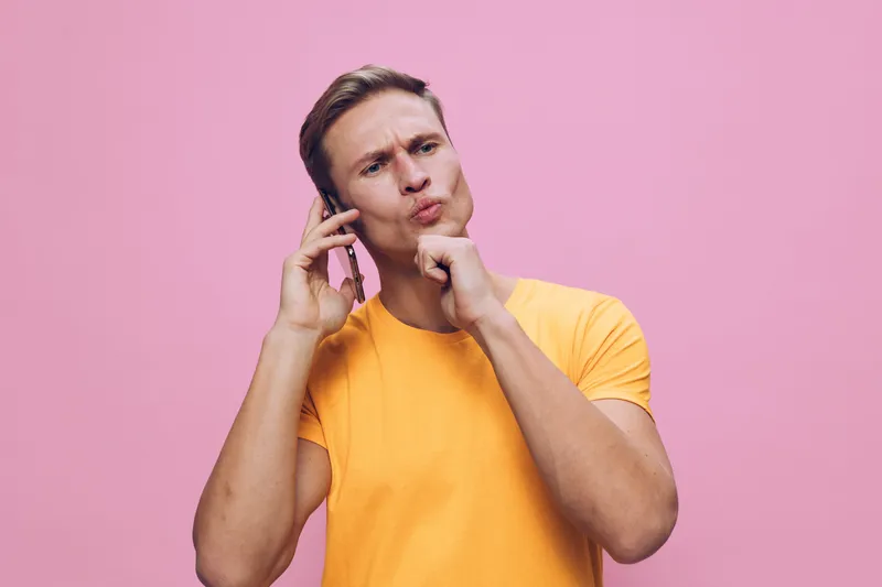 pensive young caucasian man talking phone hand on chin wear orange t-shirt pink background digital nomad concept communication technology