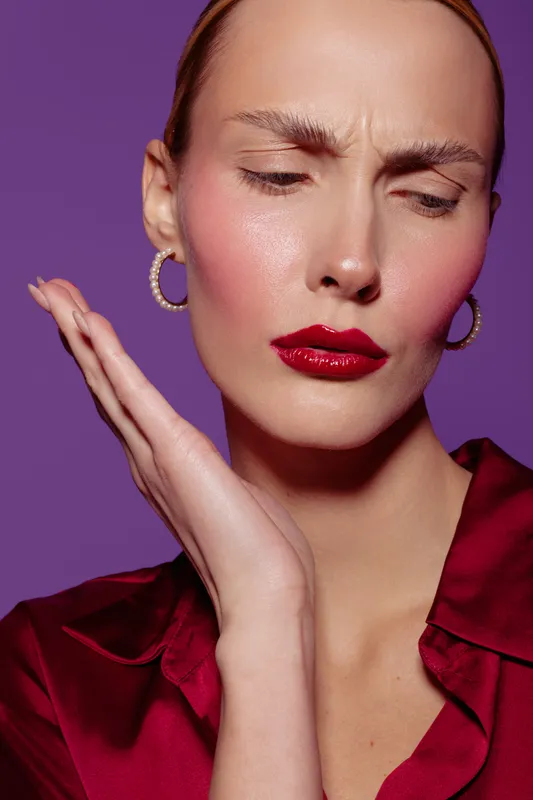 Close-up of a woman with red lipstick and hoop earrings wearing a red satin blouse, posing with a thoughtful expression on a purple background. Concept of beauty and fashion.