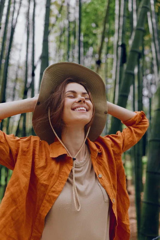 Woman smiling in hat and orange shirt in bamboo forest, portrait of happiness and nature. Outdoors travel scene with relaxed pose, green grove background and peaceful mood.