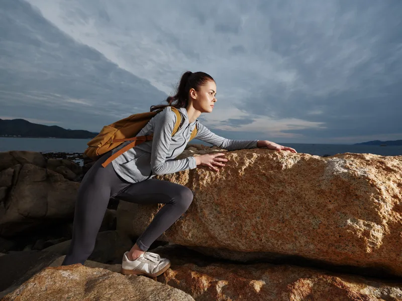 Active young woman exploring rocky coast, wearing athletic clothing and backpack, showcasing determination against dramatic background