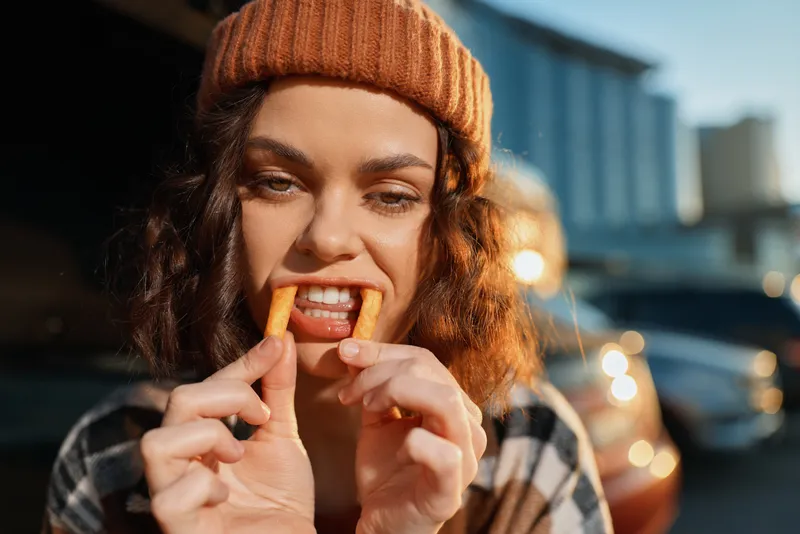 Woman portrait fries candid beanie urban smile golden hour authenticity playful mood, candid lifestyle and emotional storytelling of a young woman eating a snack outdoors in warm glow.