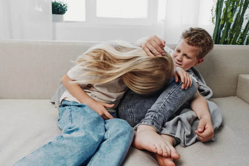 Two playful children sitting on a couch in a bright room, a boy and a girl laughing and teasing each other, casual clothes and natural light create a warm, joyful atmosphere.