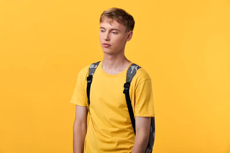 Young man in a yellow t shirt with a backpack stands against a bright yellow backdrop, casual style, relaxed mood, everyday fashion, minimal setup, suitable for lifestyle, education, travel and youth