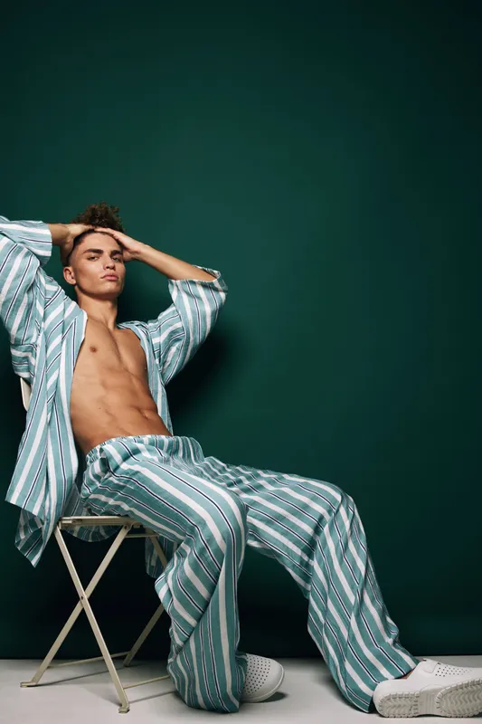 Young man in striped pajamas sitting on chair with hands behind head, relaxed pose and casual homewear against dark green background in studio.