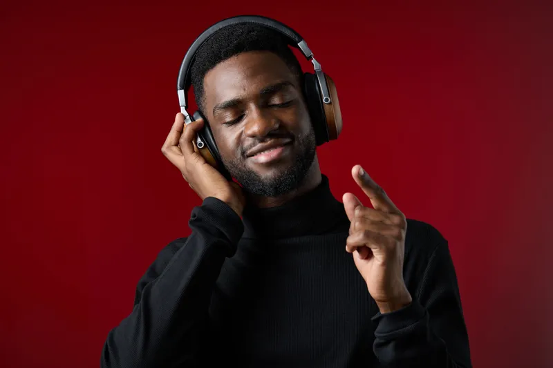 Portrait of a smiling young Black man enjoying music with headphones, wearing a stylish black turtleneck, set against a rich red background