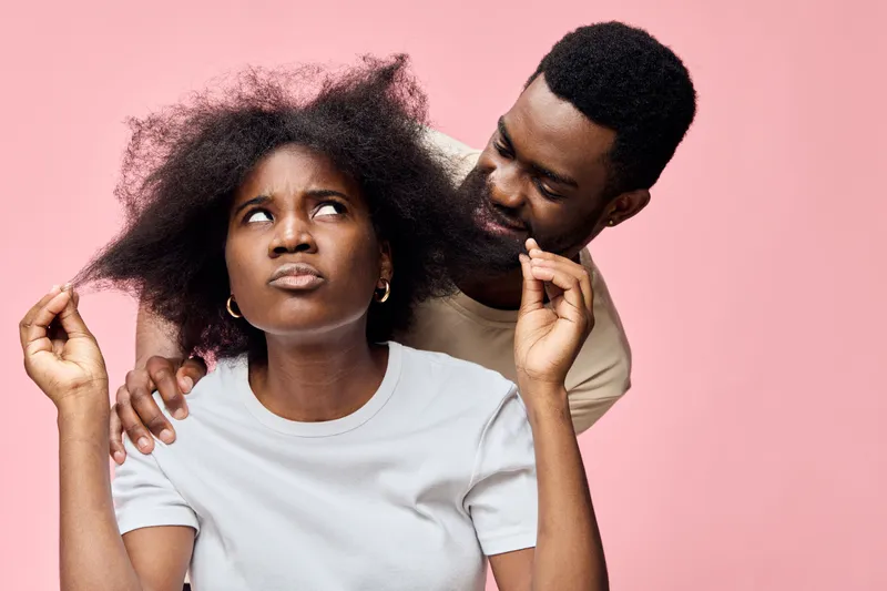 A young African couple expresses frustration and curiosity, with the woman showing concern about her hair while the man looks on with interest, set against a soft pink background