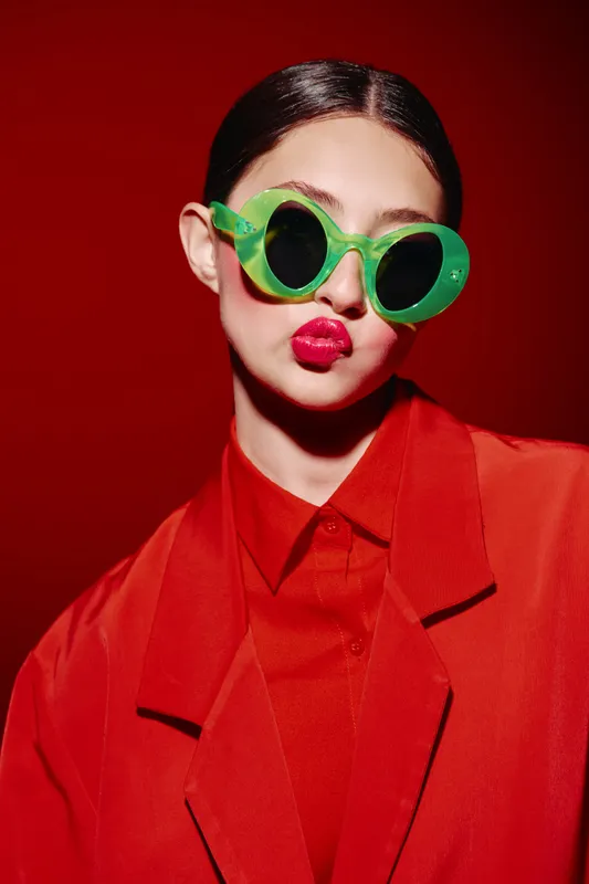 Stylish woman in vibrant retro green sunglasses and bright red outfit poses on red background. Bold makeup and confident expression enhance fashion beauty concept.