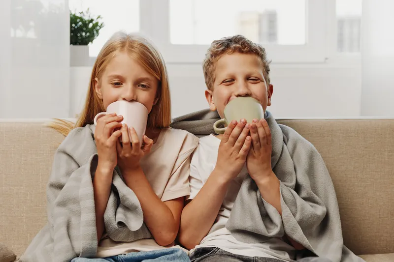 Two children wrapped in gray blankets enjoy hot drinks at home, sitting on a beige sofa near the window. Cozy atmosphere and warmth concept.