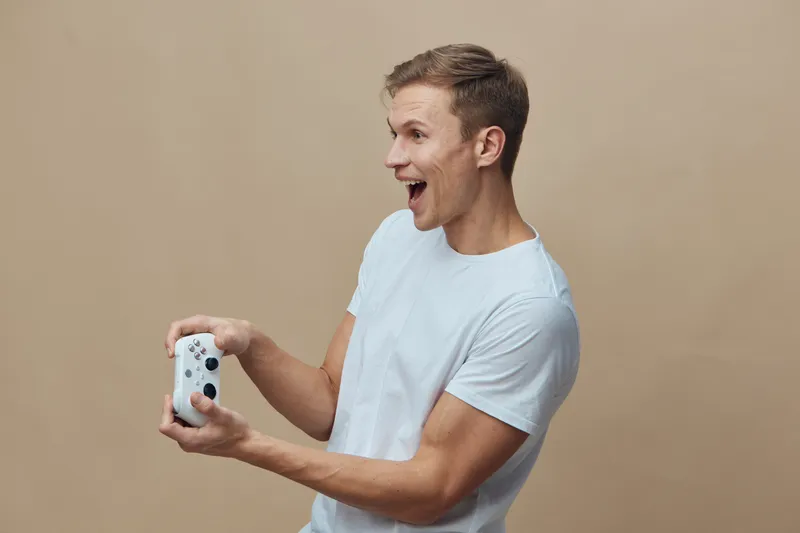 Smiling young man with short brown hair wearing casual white t-shirt holding a gaming console with one hand isolated on beige background studio portrait. People lifestyle concept