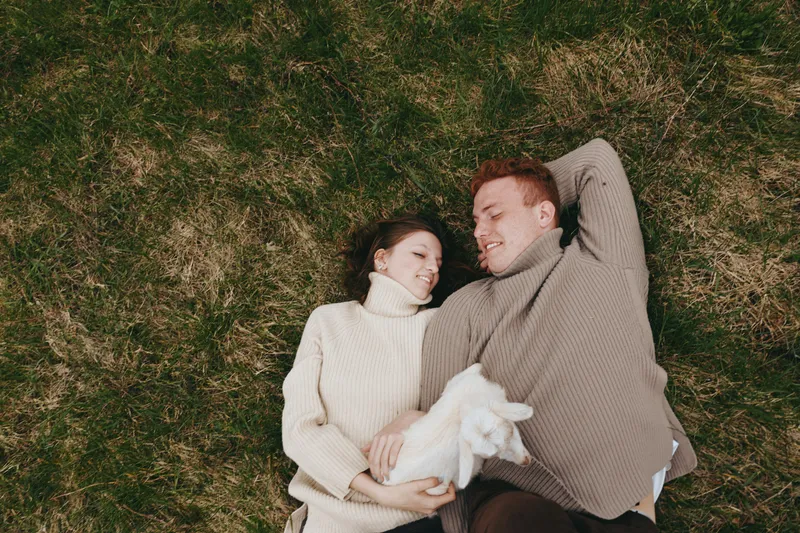 A couple lies on a grassy meadow, cuddling a small goat kid between them. The scene conveys relaxed lifestyle, outdoor bonding, and gentle affection in a natural farm setting.