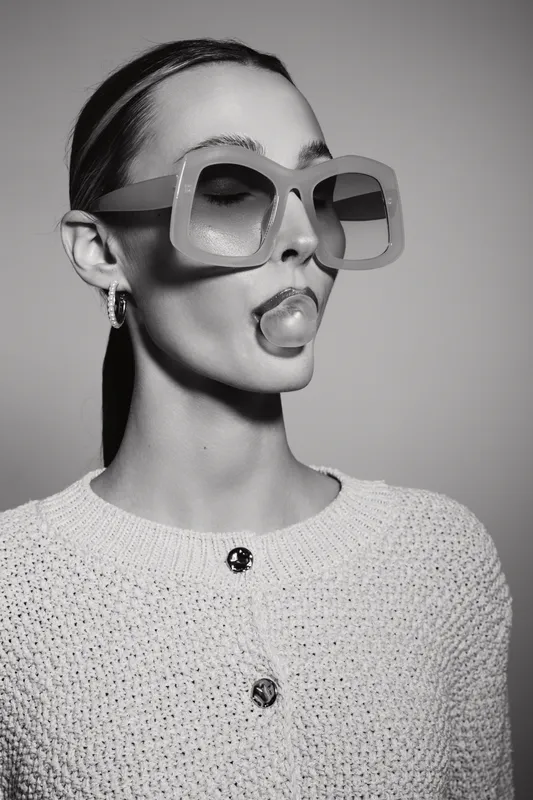 Young caucasian woman with pale skin wearing oversized sunglasses and blowing bubble gum, posing in knitted sweater in studio.