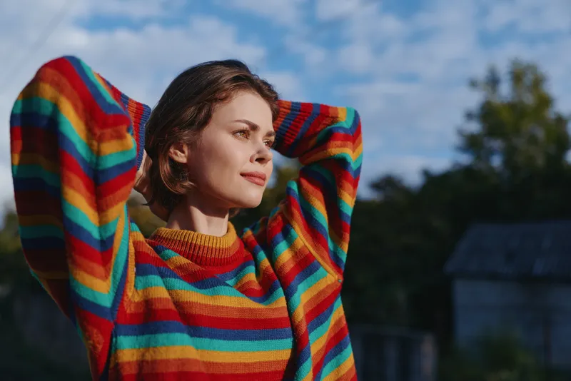 Young woman striped sweater outdoors relaxed smiling casual outfit nature countryside with hands behind head, colorful fashion portrait in soft natural light for lifestyle travel.