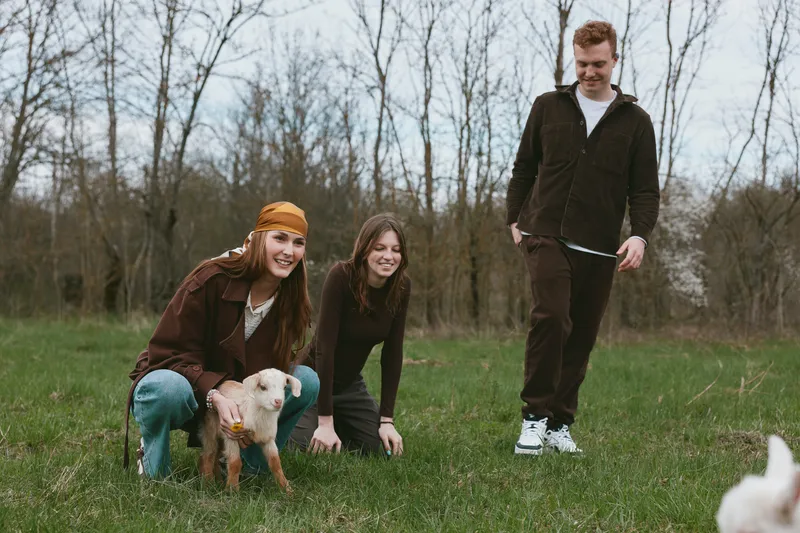 Casual scene of three friends outdoors with a dog in a grassy park, candid smiles, warm jackets, relaxed lifestyle moment, natural light creating a cheerful mood, everyday day out