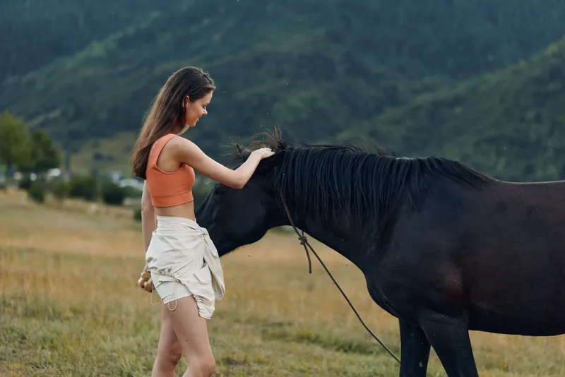A relaxed woman in casual attire stands beside a calm horse in a wide meadow, gently reaching to pet its neck. The tranquil countryside setting conveys connection, care, and a peaceful outdoor